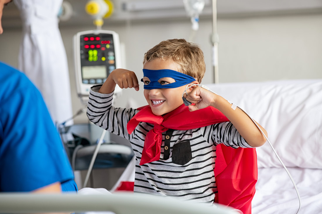 Young boy wearing superhero costume in hospital bed and smiling while making superhero pose.